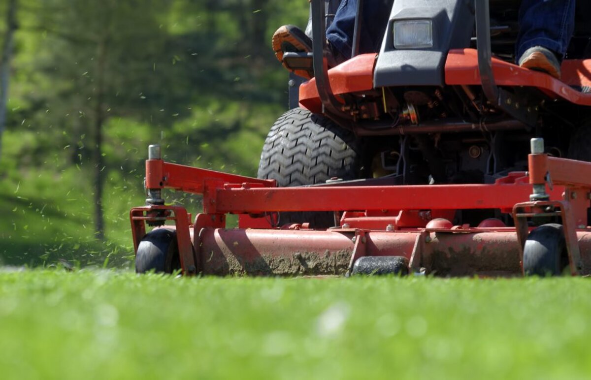 Close-up of a commercial mower deck cutting green grass with clippings spraying out — the kind of mowing that continues into fall until the grass stops growing
